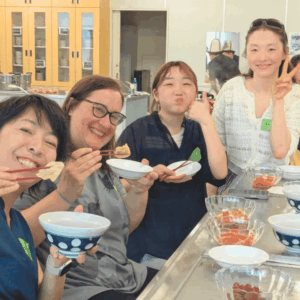 食事を楽しみながら、カメラに向かってポーズをとる、日本、メキシコ、スイス、モンゴル出身の女性４人の様子