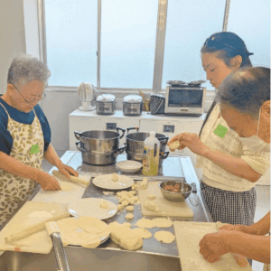 年配の日本人女性１人と年配の日本人男性１人と、モンゴル出身の女性が一緒にモンゴルの家庭料理、包子（ヴォーズ）を包んでいる様子