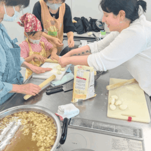 日本人の小学生女の子が、ドイツ出身の女性から、モンゴルの家庭料理、包子（ヴォーズ）の包み方を教えてもらっていて、それを見守る日本人女性２人の様子