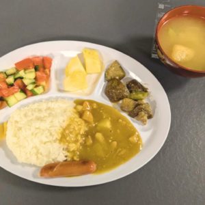 ネオアス村地域食堂で提供されたカレーやサラダ、ナスの炒め物、パイナップルなどが盛り付けられているプレートと、お味噌汁の写真