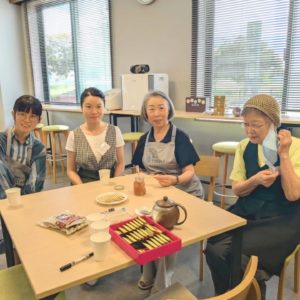 ネオアス村地域食堂の4名の女性ボランティアスタッフが、椅子に座ってお茶をしながらカメラに向かって笑顔を見せている様子